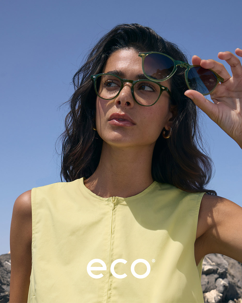 Eco Eyewear campaign featuring female model in green Biobased eyeglasses holding the matching sun clip-on in outdoor settings – sustainable women’s eyewear fashion photography.