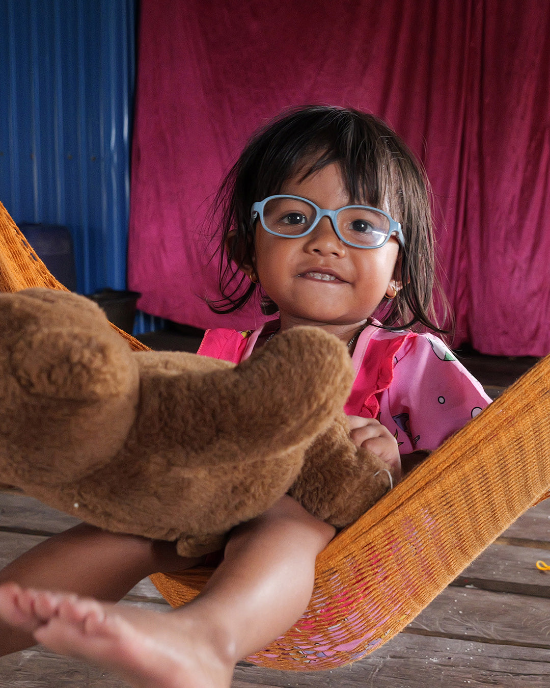 Young child wearing light blue kids’ eyeglasses, sitting on a hammock and holding a teddy bear.