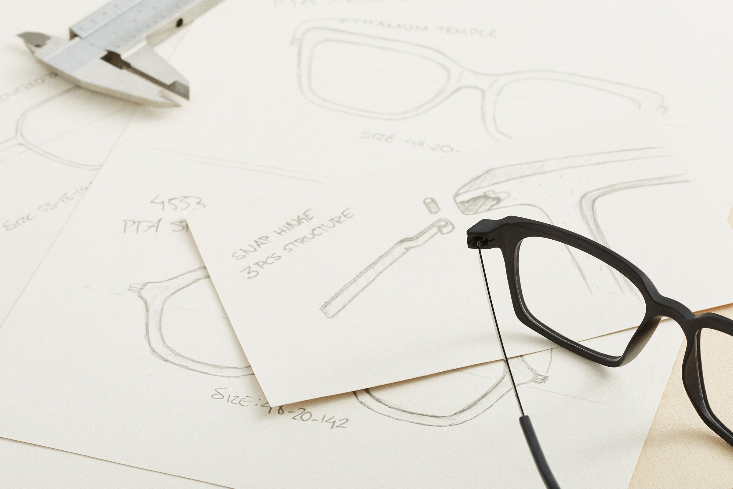 Flatlay of eyeglass design sketches with a black eyeglass frame and a vernier caliper placed on top.