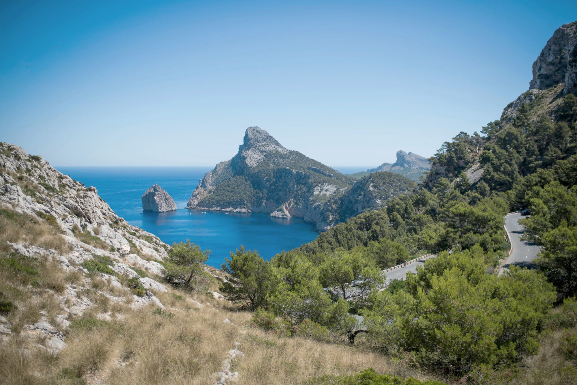 Bright coastal landscape with sea, green hills, and a winding road—viewed with the natural eye.